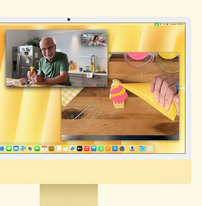 iMac amarelo. Demonstração da Vista de Secretária. O ecrã mostra uma pessoa numa pastelaria a fazer uma videochamda com outra pessoa. Um dos ecrãs mostra as pessoas que estão a fazer a videochamada e o outro ecrã mostra as mãos da pessoa que está a fazer a cobertura de uma bolacha