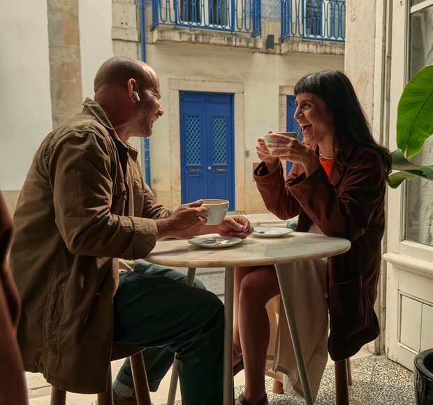 A person wearing AirPods Pro 3, laughing in conversation with another person, both holding a cup of coffee