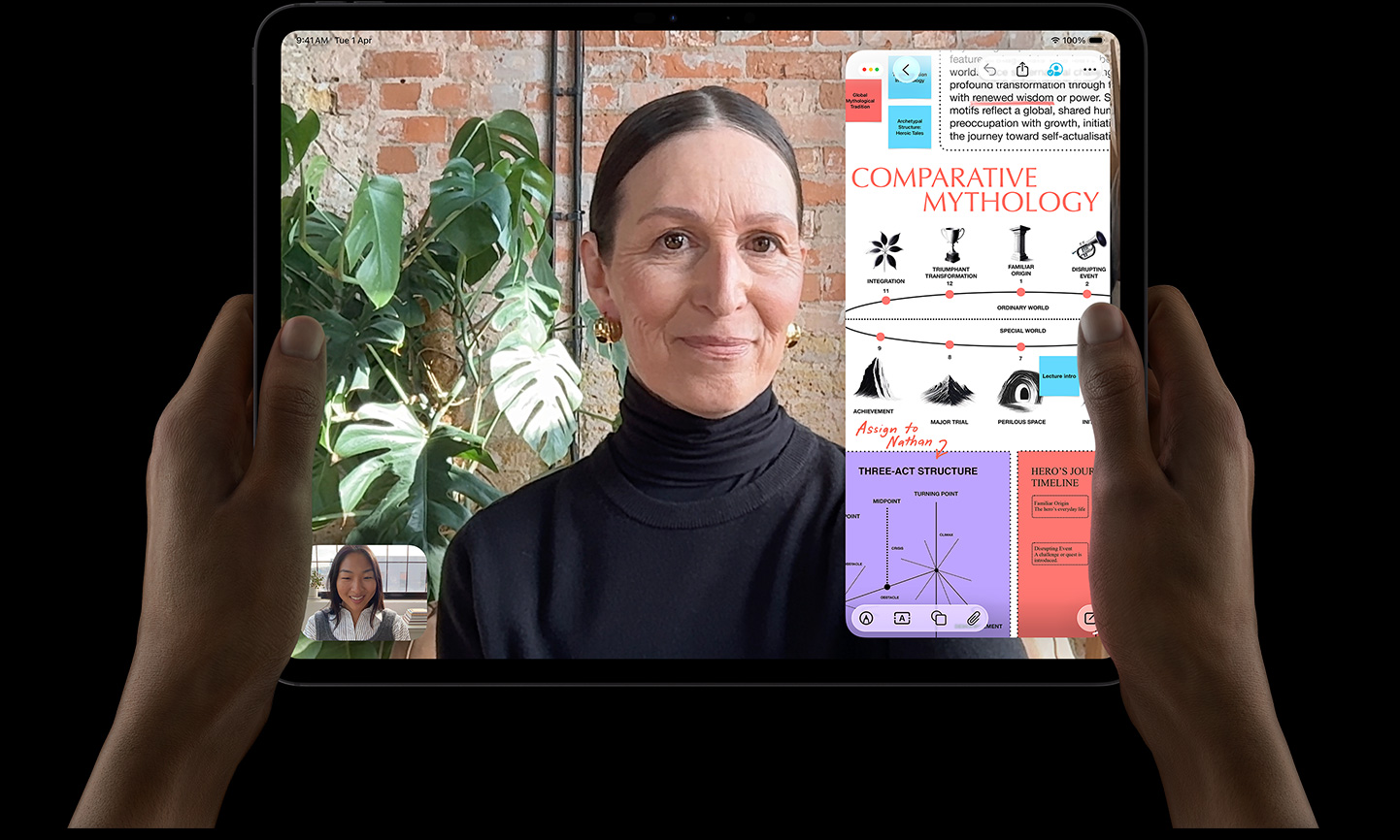 iPad Pro, front exterior, space black colour, left and right hands holding in landscape orientation, rounded corners, black display bezel, display showing a FaceTime conversation being live translated and an educational app showing a presentation of 'Comparative Mythology' with text, diagrams, and handwritten notes