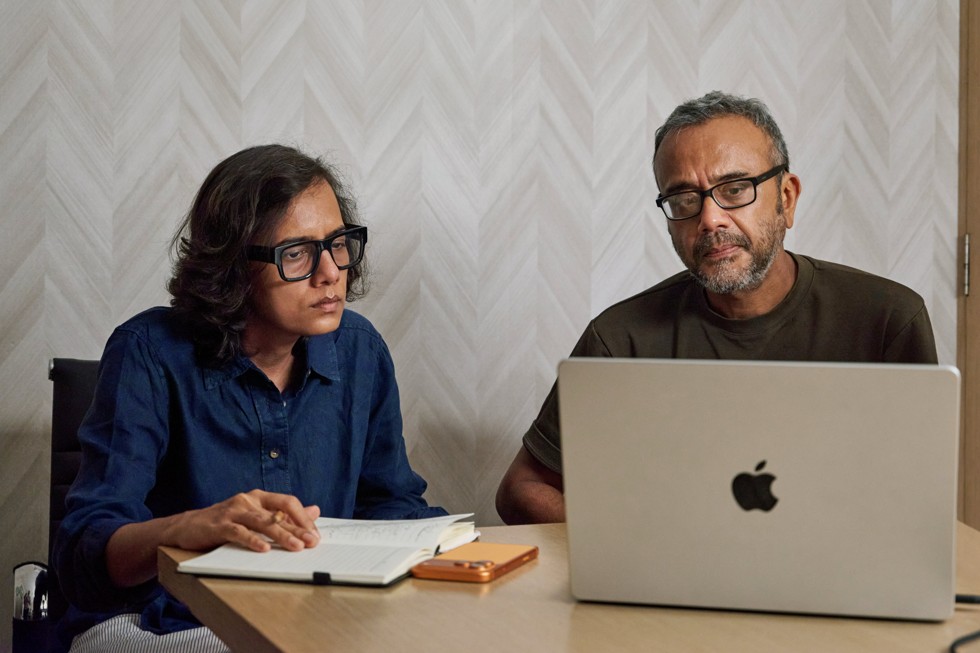 Dibakar Banerjee e Dhritisree Sarkar estão sentados à mesa, olhando para um MacBook Pro. Um iPhone 17 Pro Max está sobre a mesa ao lado deles.