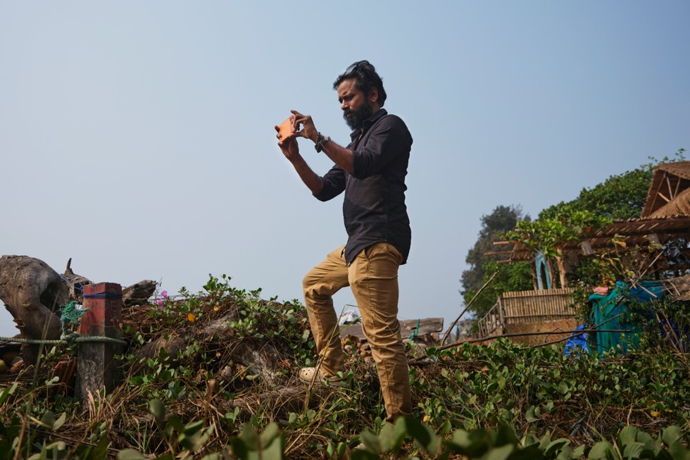 Ritesh Sharma está em uma colina gramada com vista para a água, segurando o iPhone 17 Pro Max enquanto filma.