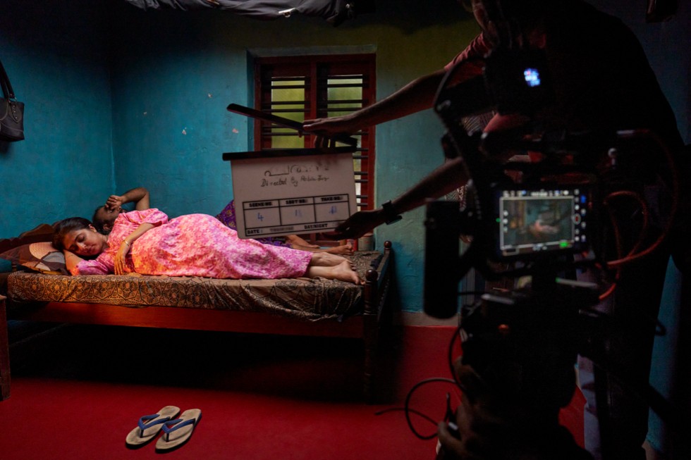 Um set de filmagem mostra uma pessoa deitada em uma cama em um ambiente com pouca luz, com equipamentos de câmera visíveis.
