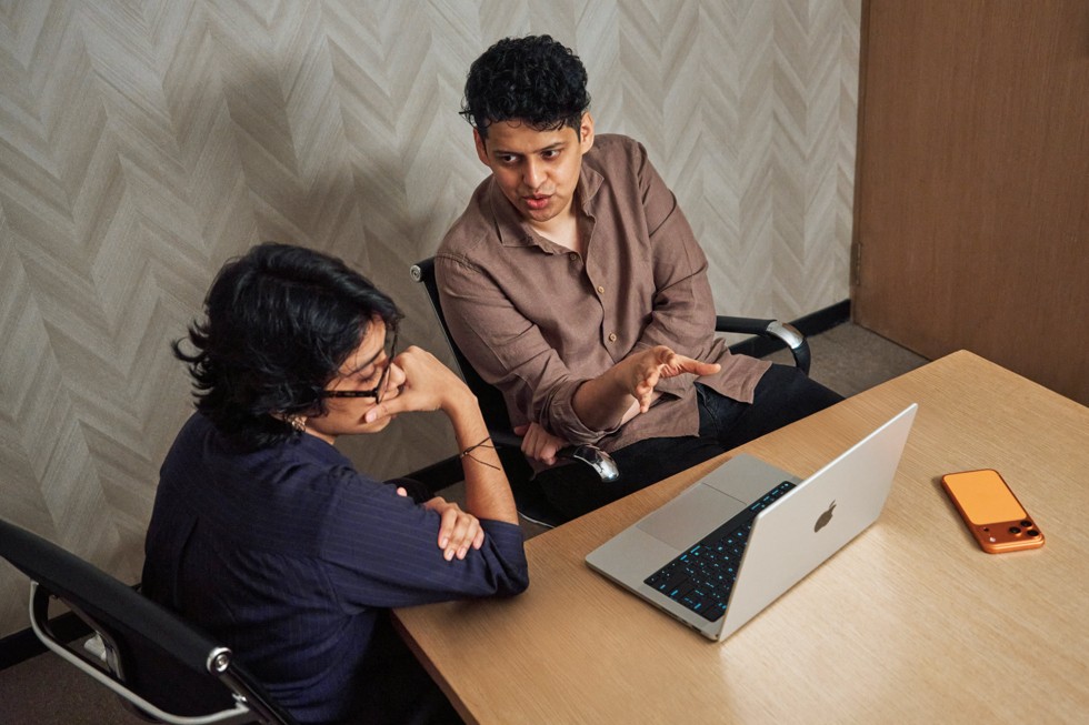 Shreela Agarwal está sentada à mesa com o mentor Chaitanya Tamhane, olhando para um MacBook Pro e conversando. Um iPhone 17 Pro Max está sobre a mesa ao lado deles.