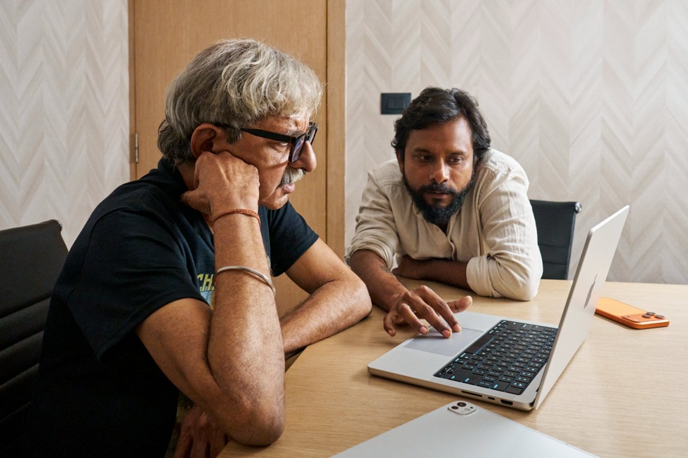 Ritesh Sharma colabora com outro integrante da equipe em uma mesa, olhando para um MacBook Pro. O iPhone 17 Pro Max está sobre a mesa ao lado deles.