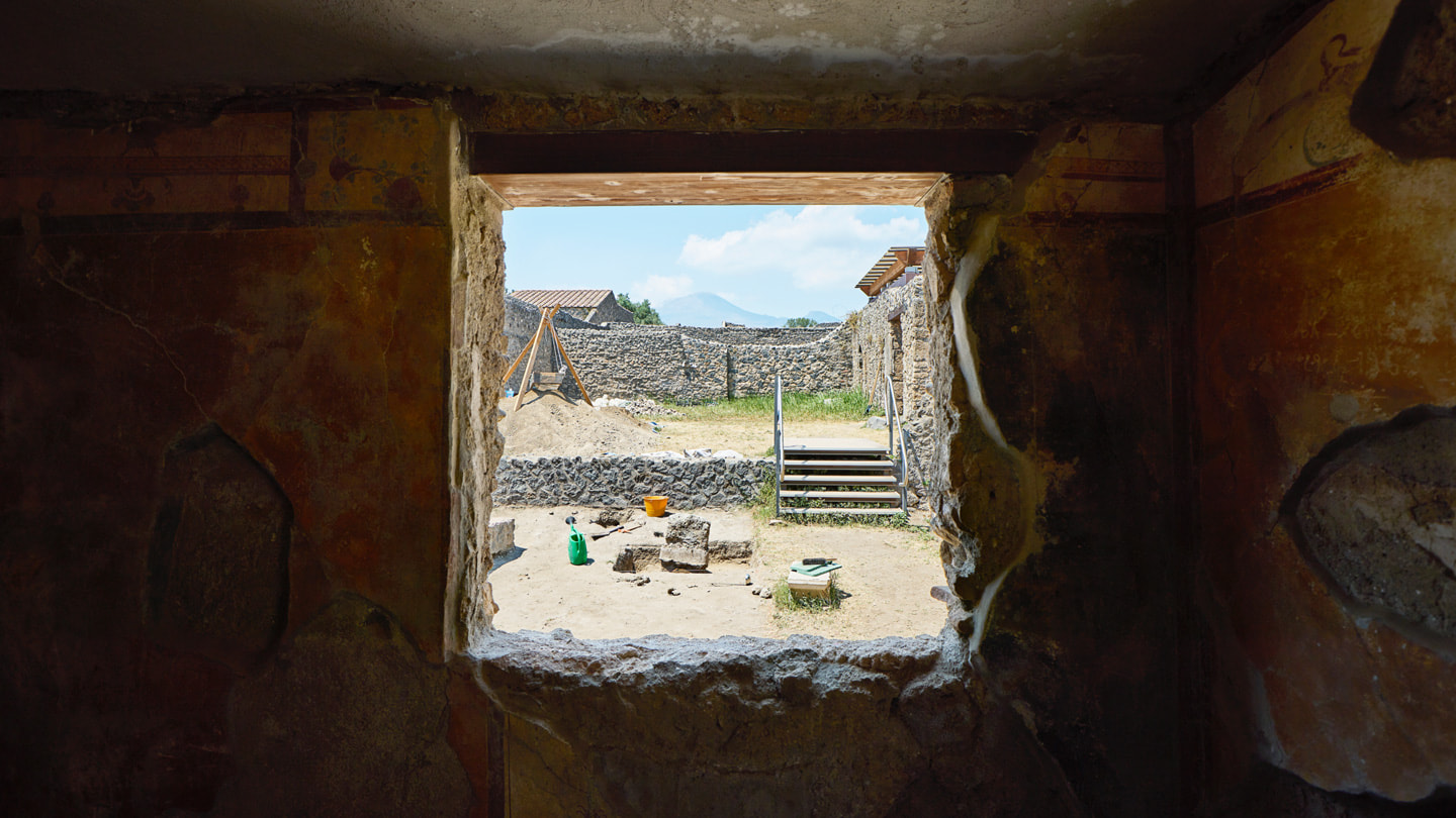 Der Blick durch ein Fenster in der Ausgrabungsstätte zeigt Mauern aus Steinen und den Vesuv in der Ferne. 
 