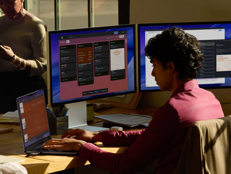 Equipe de funcionários sentada em torno de uma mesa em um escritório. Uma mulher sentada à mesa trabalha em um MacBook e em monitores. Colegas se reúnem para uma discussão