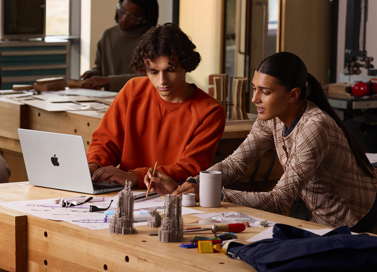 Duas pessoas a trabalharem num projeto numa mesa, uma a digitar no MacBook e a outra a usar um lápis e uma régua num papel, rodeadas de material de arte.