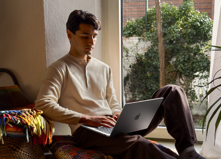 Uma pessoa sentada no chão a usar o MacBook junto a uma janela.