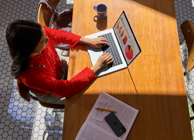 Uma pessoa a usar o MacBook numa mesa, com uma caneca, smartphone e papéis por perto.