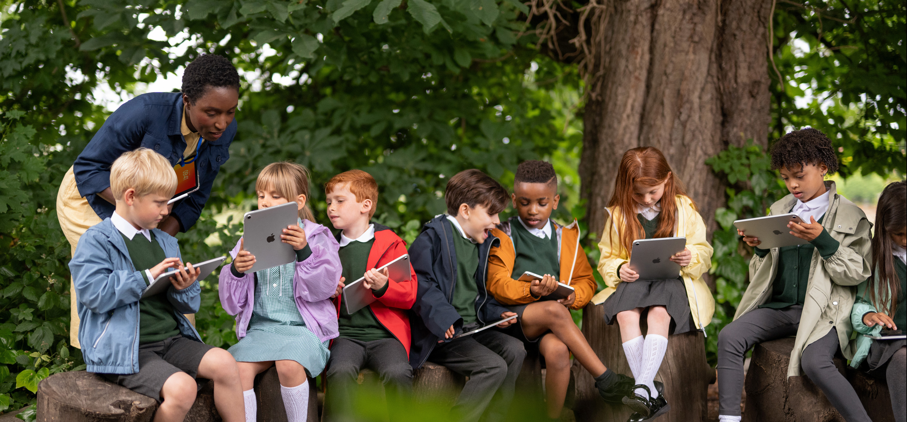 Un grupo de estudiantes de primaria usa un iPad al aire libre bajo la supervisión de una docente, que también sostiene un iPad, se destaca la facilidad de aprender en cualquier lugar