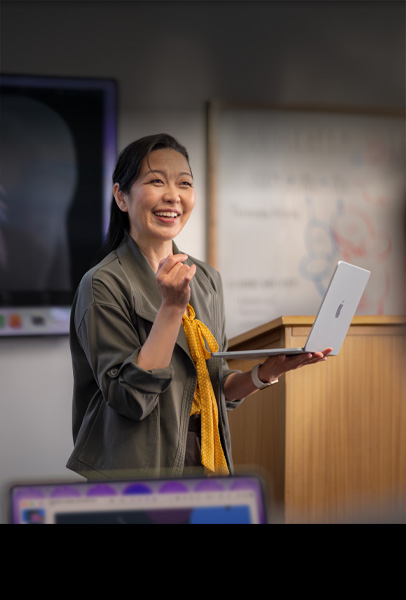 Una profesora hace una presentación a sus estudiantes en un salón mientras sostiene una MacBook color plata, se muestra el rendimiento y la larga duración de la batería
