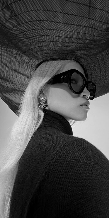 Gros plan en noir et blanc d’une personne avec de grandes lunettes de soleil et un grand chapeau qui sort du cadre.