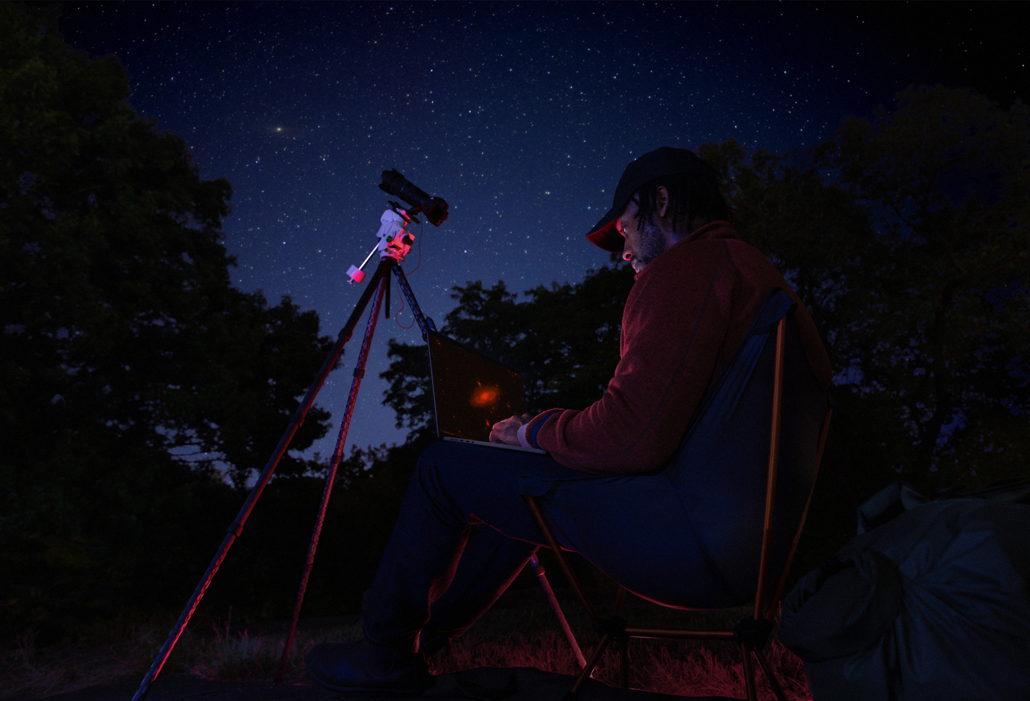 Una persona que está al aire libre, de noche, tiene una MacBook Pro apoyada en su regazo conectada a un dispositivo con un trípode que apunta hacia las estrellas
